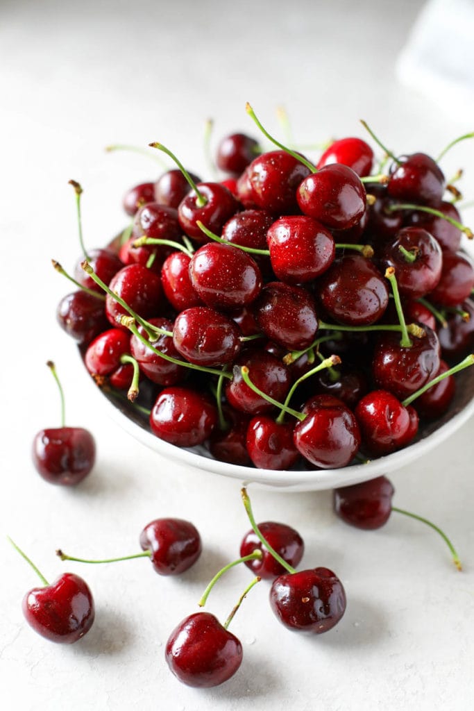 Bowl of sweet cherries with stems in a white bowl surrounded by scatter whole cherries.