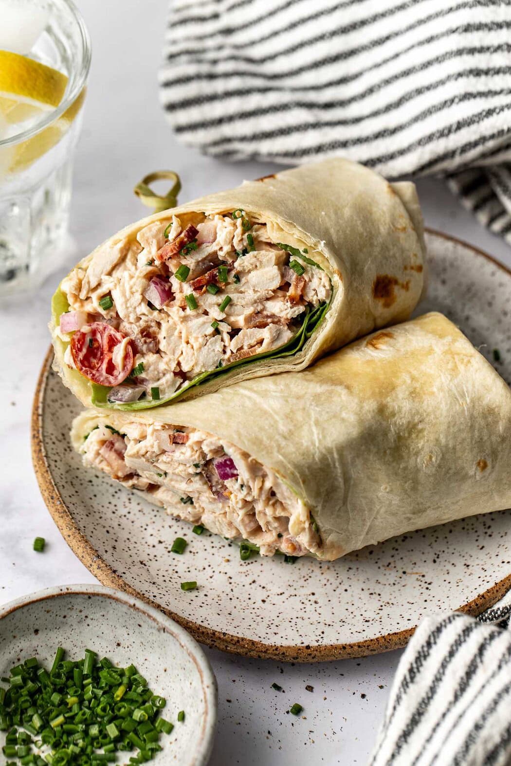 Close up view of a plate with a BLT Chicken Salad wrap on it next to a small bowl of chives. 