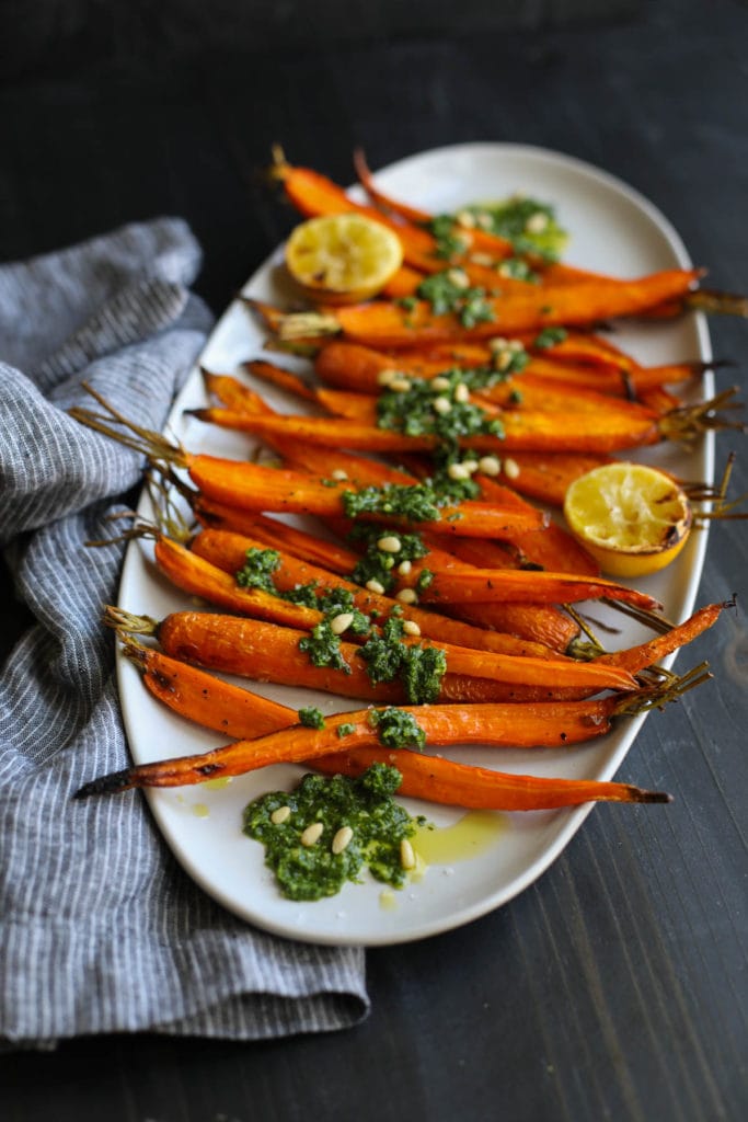 Roasted carrots on a white platter topped with pesto and roasted lemon wedges.
