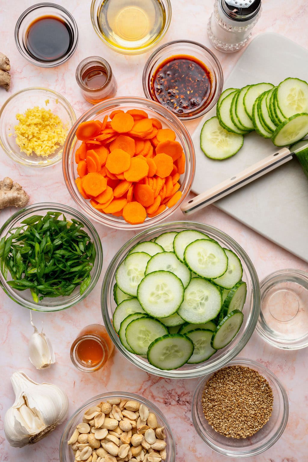 Overhead view of a variety of ingredients for Asian Cucumber Salad in different sized bowls on a white marble countertop.