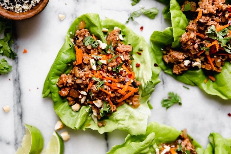 Close up view of a lettuce wrap containing Chinese-inspired pork topped with shredded carrots and fresh herbs. 