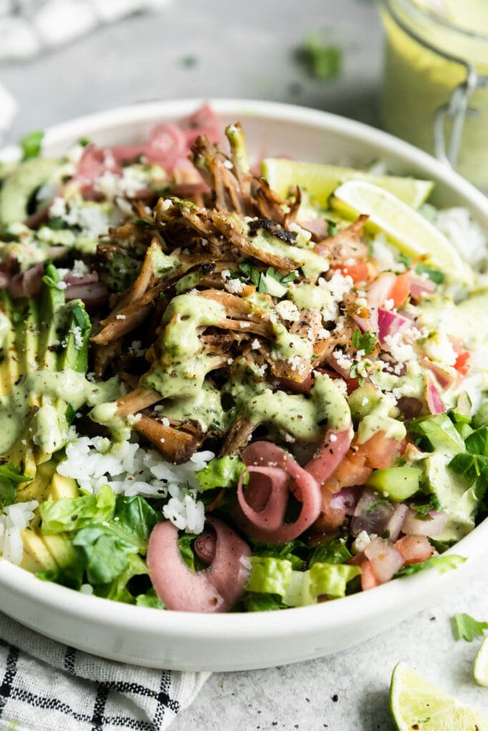 Close up view of carnitas burrito bowl in a white bowl filled with white rice, slow cooker carnitas, pink pickled onions, lime wedges, greens, and drizzled with cilantro-lime crema.