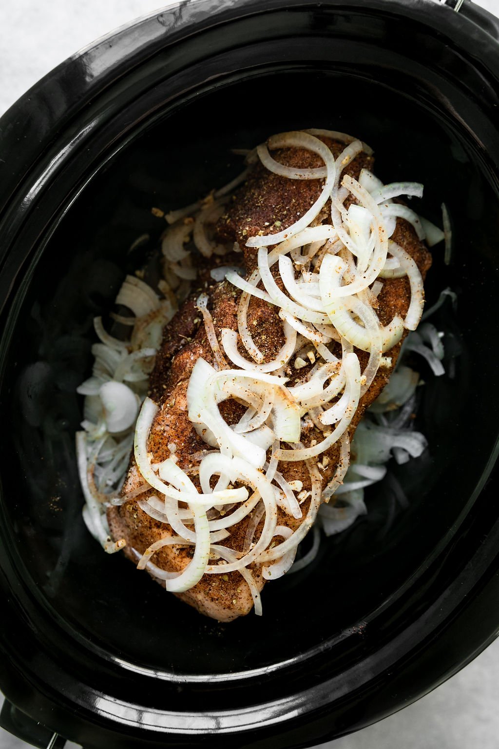 Overhead view of a pork shoulder in a black slow cooker topped with sliced onions for slow cooker carnitas.