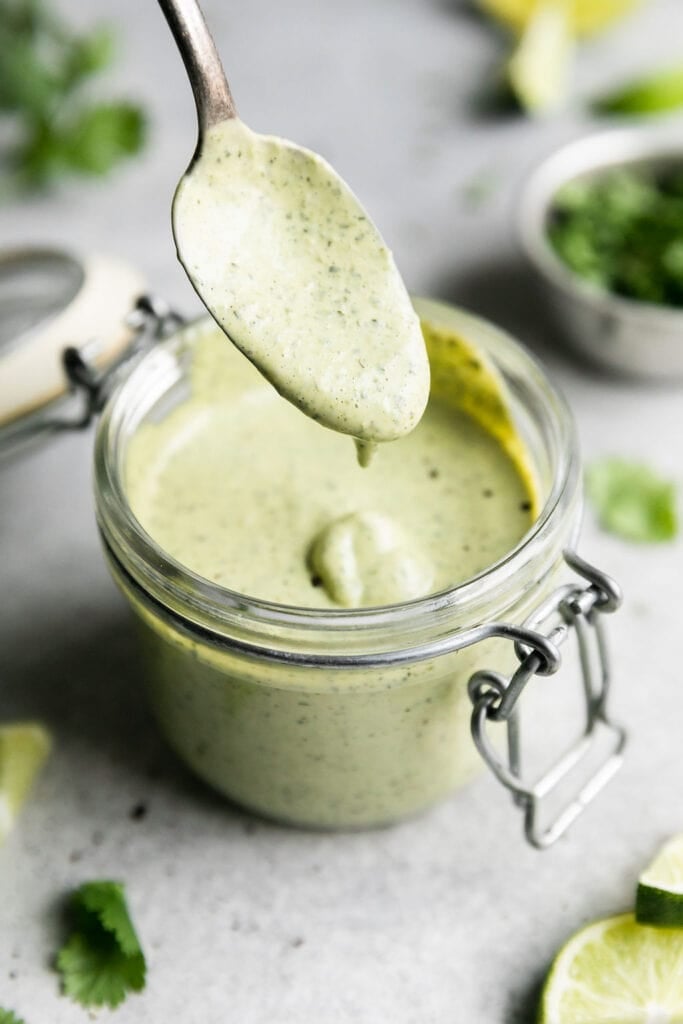 A spoon coated in creamy cilantro-lime sauce hovering over a small jar filled with green creamy cilantro-lime sauce to be drizzled over slow cooker carnitas