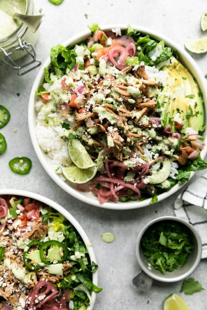 Overhead view of slow cooker carnitas in a burrito bowl with white rice, carnitas, pink onions, avocado and drizzled with avocado crema.