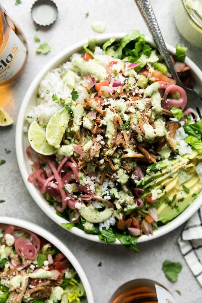 Overhead view of a white bowl filled with slow cooker carnitas, rice, pink pickled onions, avocado slices, and lime wedges for a carnitas burrito bowl