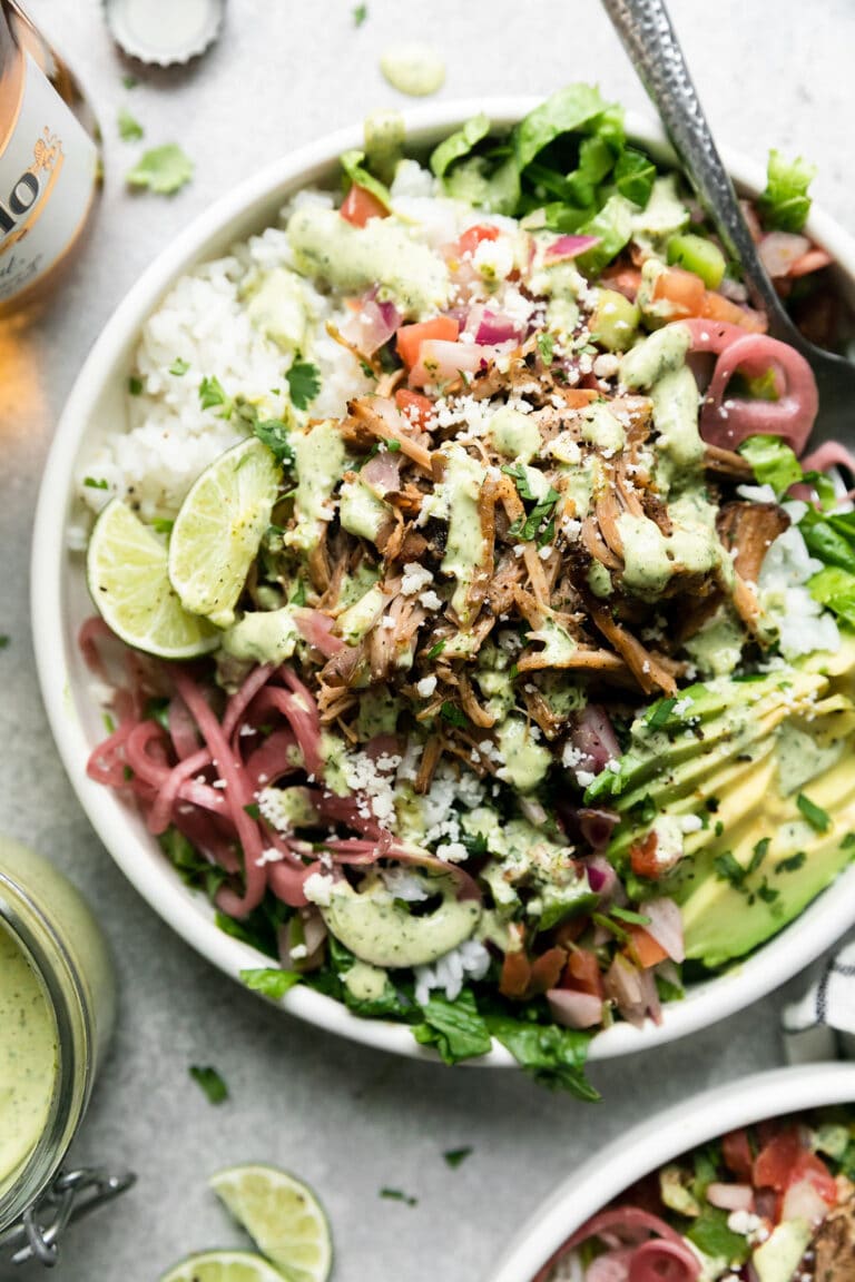 Overhead view of a plate filled with greens and carnitas topped with pickled red onions and garnished with lime wedges.