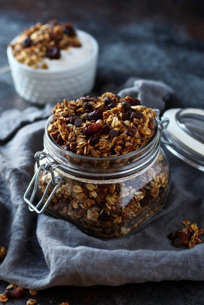 Small canning jar filled with granola surrounded by cloth napkin and a cup of yogurt with granola in the background