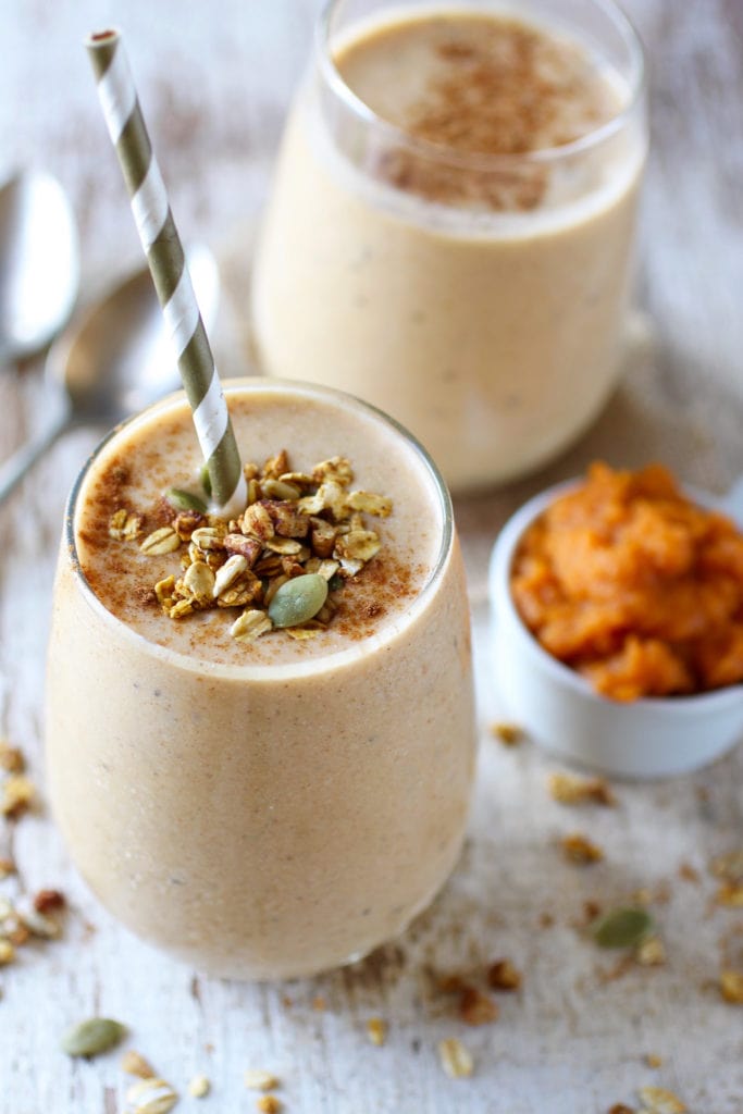 Two pumpkin smoothies in glasses, one with a straw and garnished with oats and pumpkin seeds and cinnamon with a small dish of pumpkin puree on the side 
