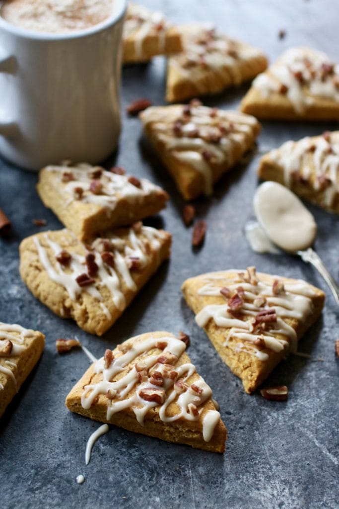 Several triangular pumpkin scones drizzled with white frosting and sprinkled with nuts with coffee cup and spoon filled with frosting 