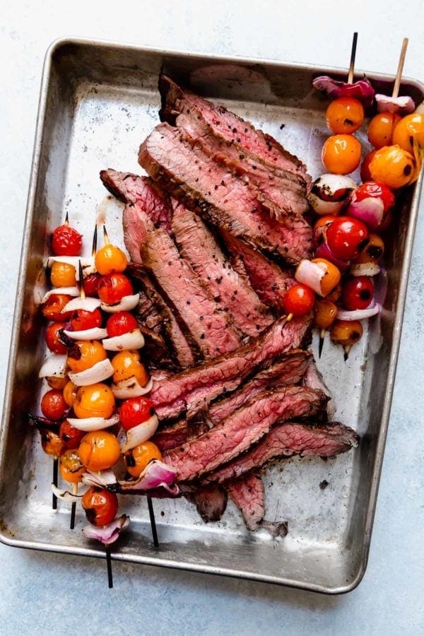 A metal baking dish filled with grilled steak sliced thin and cherry tomato and red onion kabob skewers