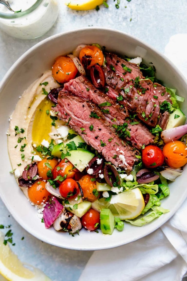 Overhead view of a grilled steak bowl with colorful vegetables and a side of hummus topped with fresh herbs. 