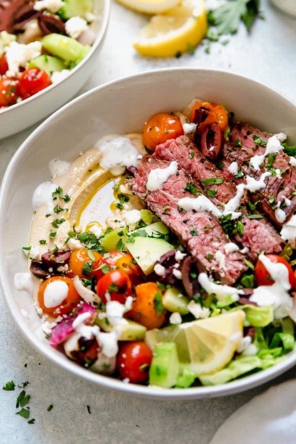In a white bowl, layers of hummus, crisp greens, grilled steak, and topped with herb-yogurt dressing.
