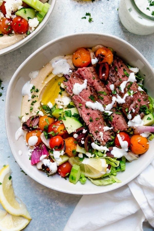 Mediterranean steak bowl with hummus in a white bowl and drizzled with herb-yogurt dressing.
