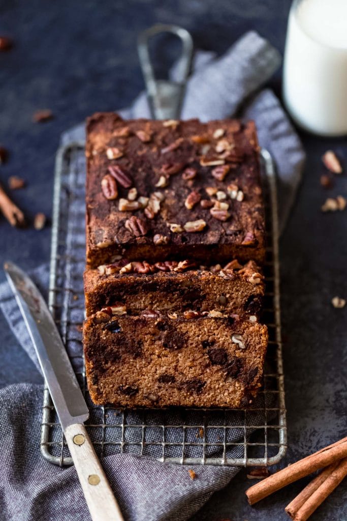Loaf of chocolate chip pumpkin bread topped with nuts resting on baking rack surrounded by glass of milk, knife and napkin