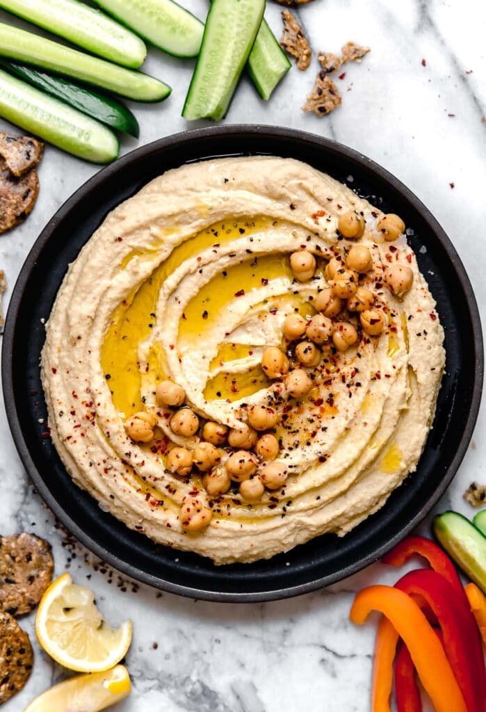 Overhead view black bowl filled with garlic hummus topped with chickpeas, olive oil, and cracked black pepper