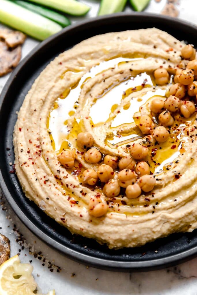 Close up view creamy garlic hummus in black bowl topped with chickpeas, olive oil, and cracked black pepper