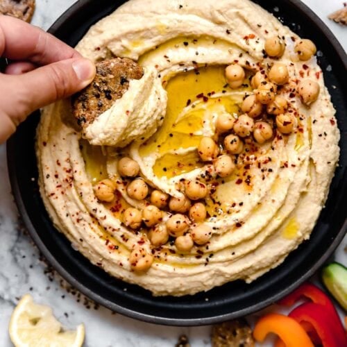 A cracker scooping up hummus from a bowl of hummus topped with roasted chickpeas and olive oil