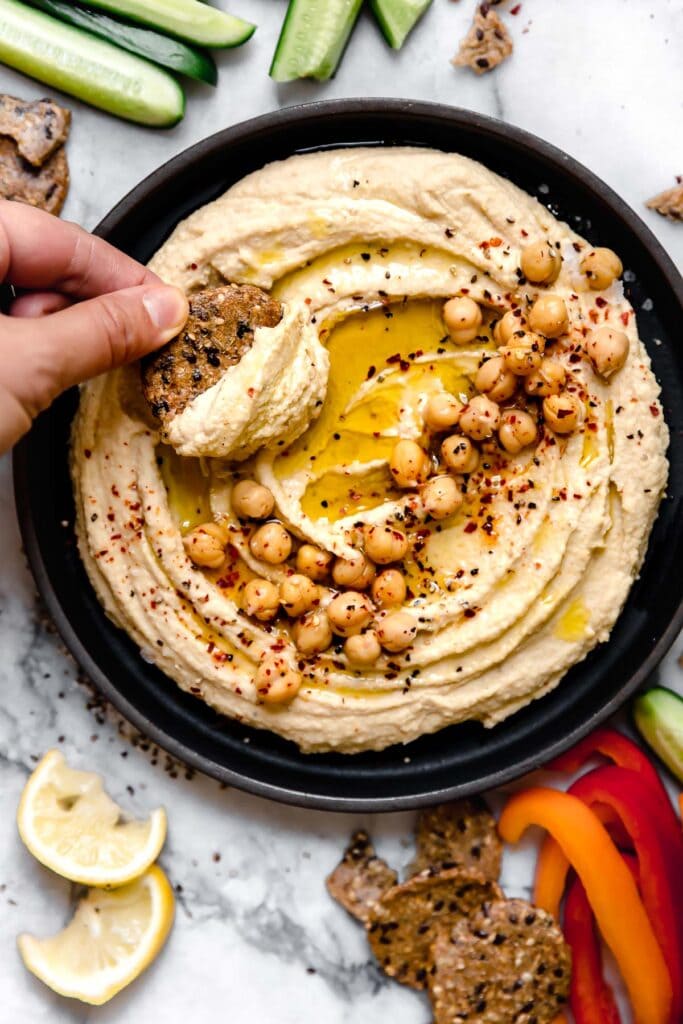 Close up view of a hand dipping a cracker into a fresh bowl of hummus topped with olive oil, chickpeas, and fresh herbs and seasonings. 