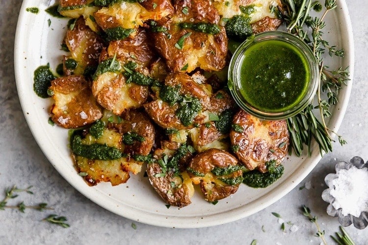 Crispy smashed potatoes stacked up on a white plate topped with green chimichuri sauce.