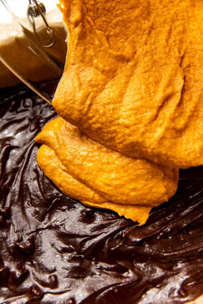 Close up view of a pumpkin cream cheese batter layer being poured over a brownie batter layer. 