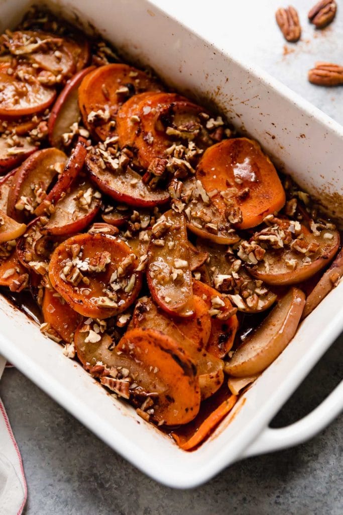 Baked sweet potato and apple slices coated in cinnamon topped with chopped pecans in white baking dish