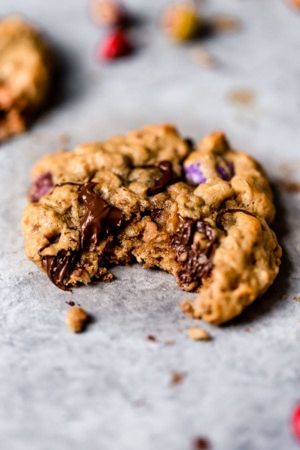 A gooey peanut butter oatmeal cookie with melty chocolate chips and colored candy pieces sits on a grey countertop with a big bite out of it. 