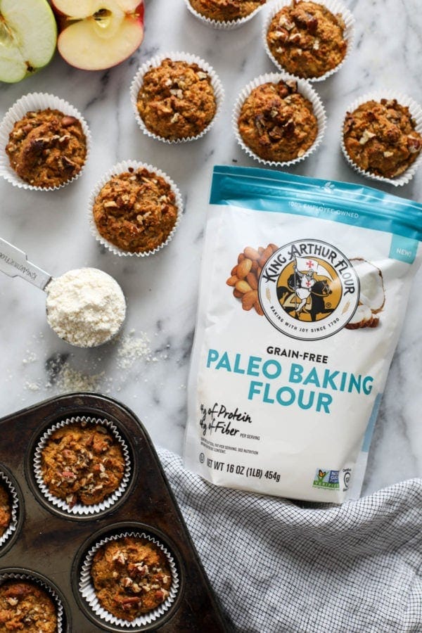 A bag of King Arthur Flour lying on a marble countertop surrounded by Paleo Morning Glory Muffins and a scoop of flour. 