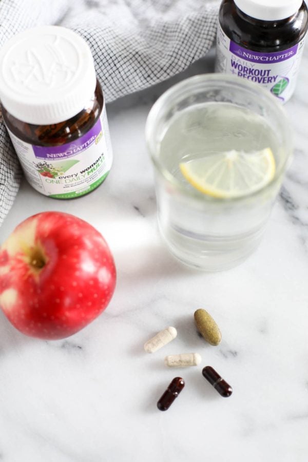 Two bottles of New Chapter supplements next to a glass of lemon water and an apple. Five tablets are on a marble countertop next to the bottles and glass. 