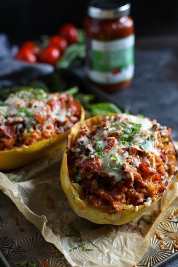 Two Pizza-Stuffed Spaghetti Squash halves on a parchment paper-lined baking sheet with ripe, red tomatoes and a jar of Primal Kitchen Tomato Basil Marinara in the background. 