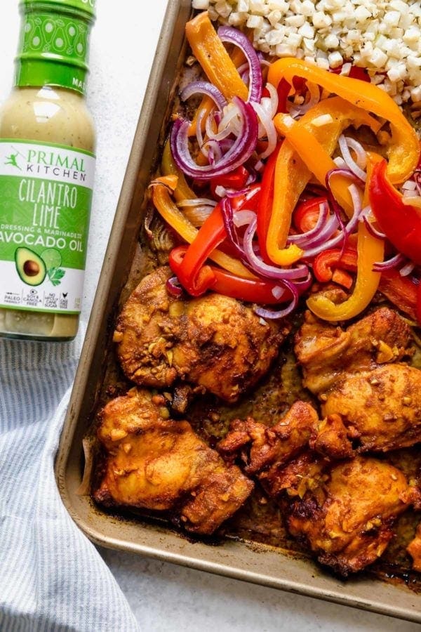 A sheet pan filled with marinated chicken thighs, bell peppers, red onions, and rice cauliflower along with a bottle of Primal Kitchen Cilantro-Lime Dressing and Marinade used to create Sheet Pan Chicken Shawarma Bowls. 