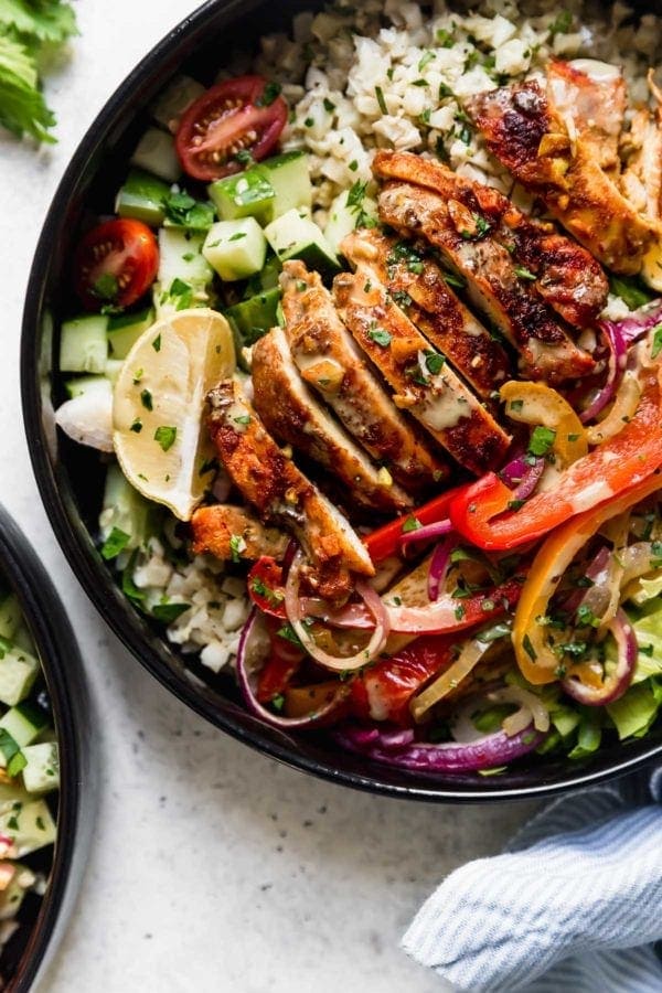 Sheet Pan Chicken Shawarma Bowls: A black bowl filled with cauliflower rice, roasted veggies, sliced chicken thighs, lettuce, cucumbers, and cherry tomatoes and drizzled with a cilantro-lime sauce. 