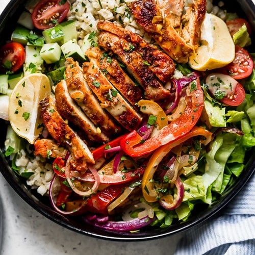 Sheet Pan Chicken Shawarma Bowls: A black bowl filled with cauliflower rice, roasted veggies, sliced chicken thighs, lettuce, cucumbers, and cherry tomatoes and drizzled with a cilantro-lime sauce.