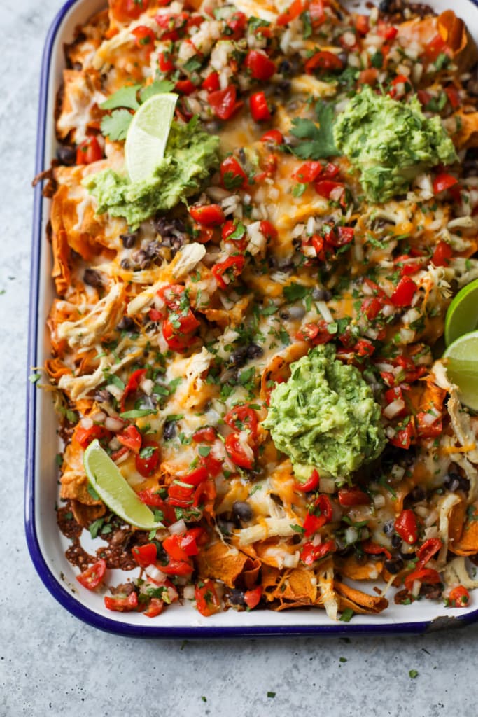 Close up view of loaded sweet potato nachos made with sweet potato chips, shredded chicken, tomatoes, melted cheddar cheese, and dollops of guacamole.