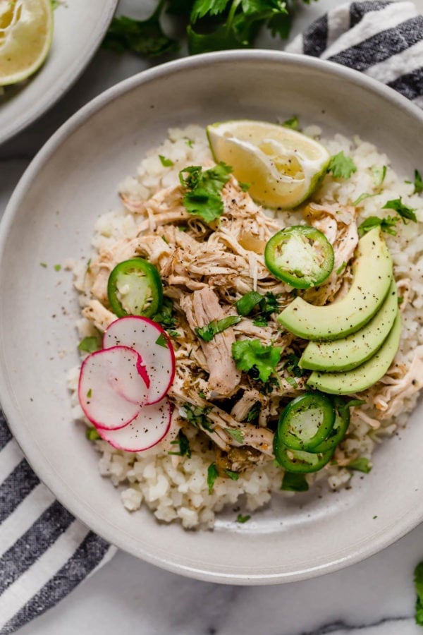 White plate on a black and white striped napkin. On the plate is a serving of Slow Cooker Chicken Chile Verde with cauliflower rice, sliced avocados and radishes, garnished with a wedge of lime.