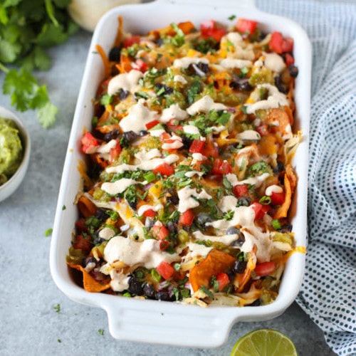 White ceramic casserole dish filled with sweet potato chips topped with melted cheese, shredded chicken, black beans, salsa, diced tomatoes, and a Southwest Ranch Dressing to create the ultimate Loaded Sweet Potato Nachos.