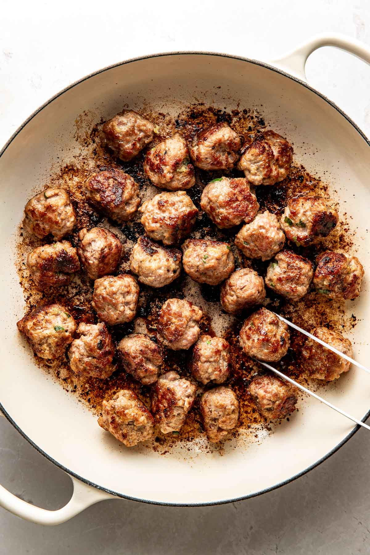 Overhead view of a white skillet filled with meat balls browning and being rotated with a tong.