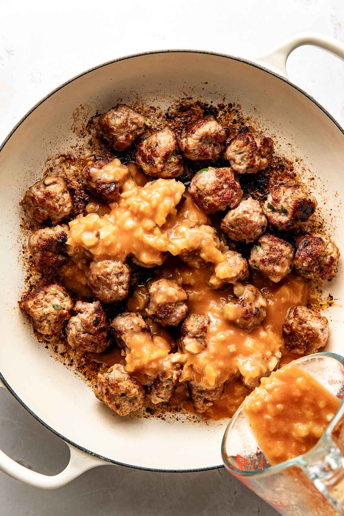 Overhead view of a white skillet filled with browned meatballs and a sweet and sour sauce mixture being poured over the top.