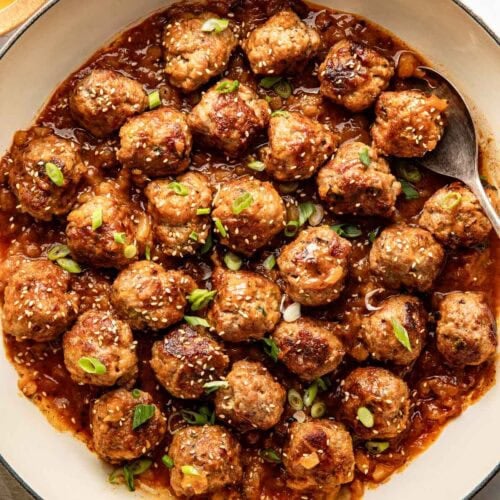 Overhead view of a white skillet filled with sweet and sour meatballs sprinkled with sesame seeds and garnished with green onions.