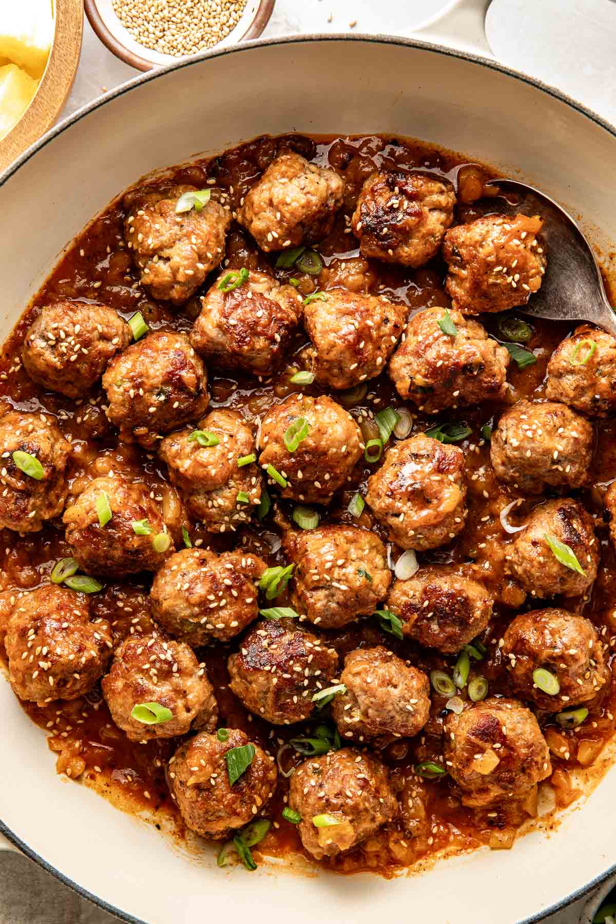 Close up view of a white skillet filled with sweet and sour meatballs sprinkled with sesame seeds and green onions. 