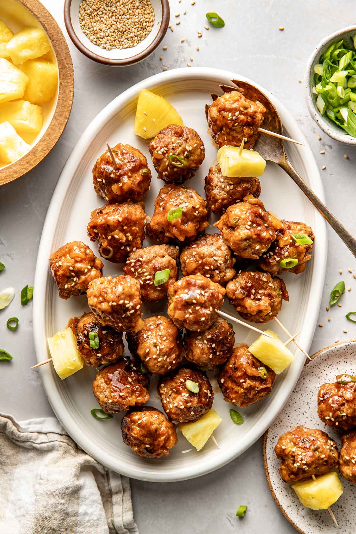Overhead view of a white oval platter filled with sweet and sour meatballs sprinkled with sesame seeds and green onions garnished with fresh pineapple pieces.