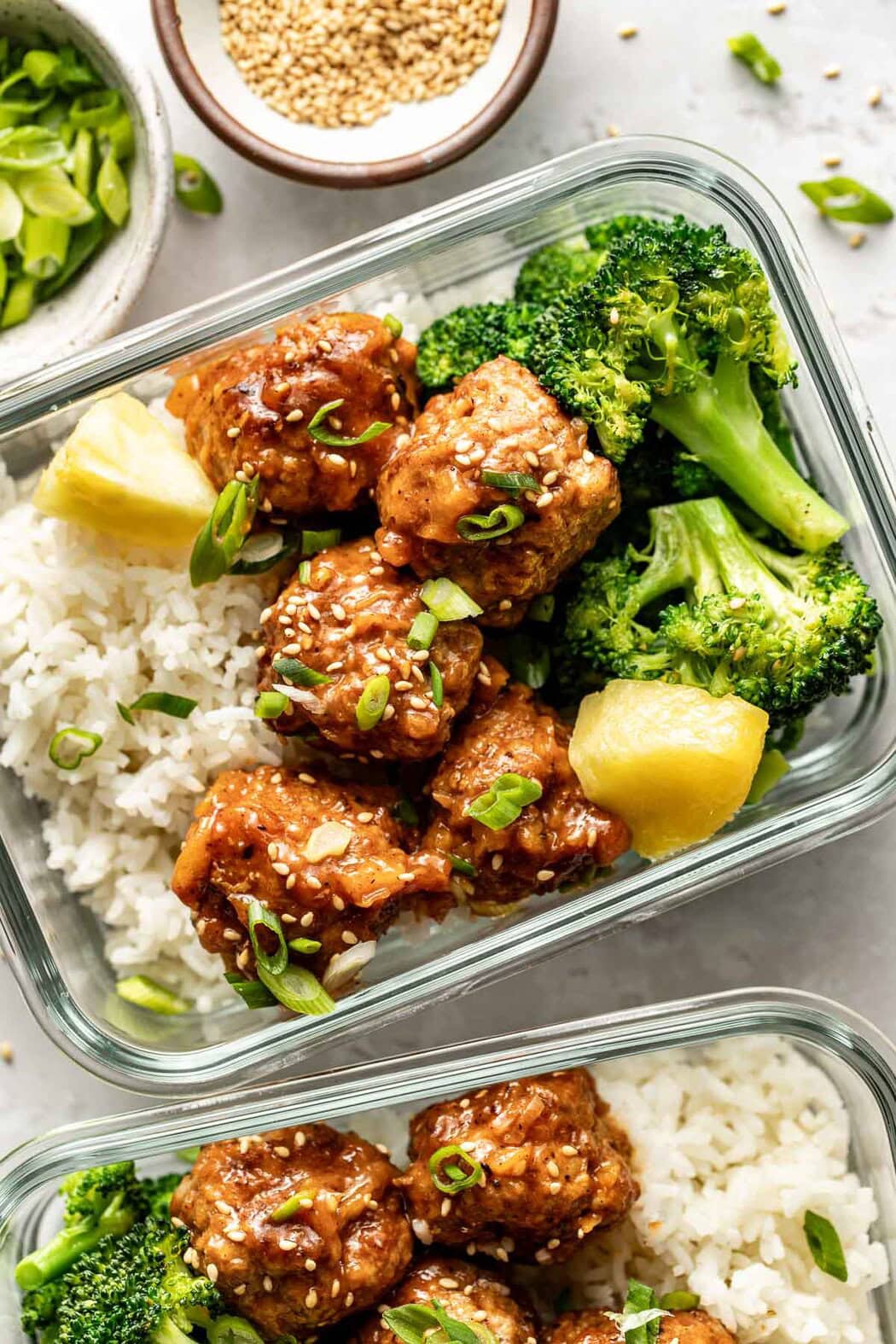 Overhead view of glass meal prep containers filled with rice, broccoli, and sweet and sour meatballs topped with fresh pineapple, sesame seeds, and green onions.