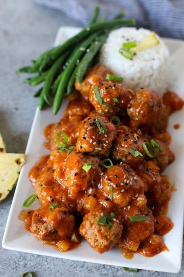 A pile of Sweet and SourA pile of Sweet and Sour Meatballs on a long white tray with steamed rice and green beans.