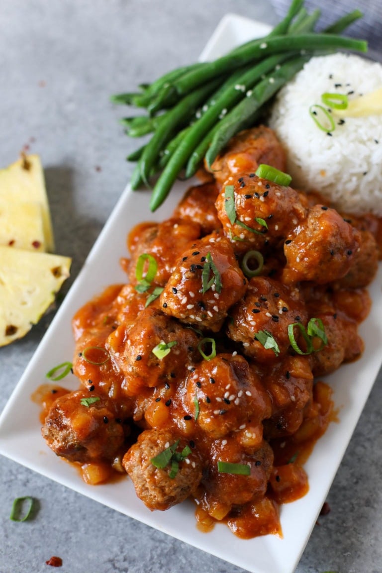Overhead view of a rectangular plate filled with sweet and sour meatballs topped with sesame seeds and rice on the side.