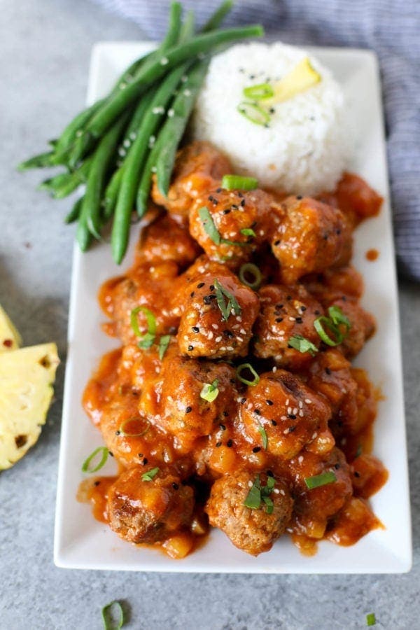 A pile of Sweet and Sour Meatballs on a long white tray with steamed rice and green beans.