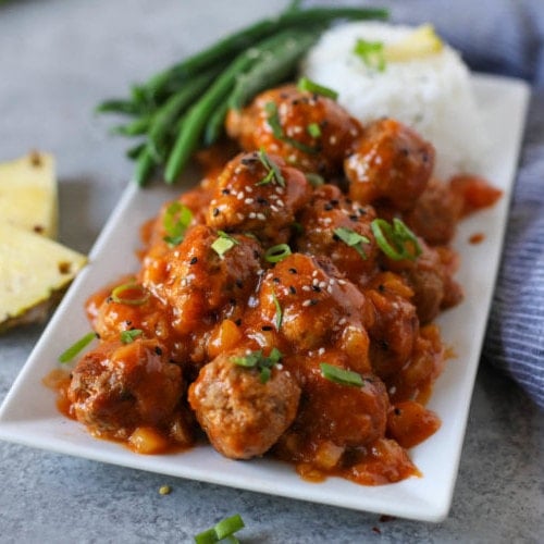 A pile of Sweet and Sour Meatballs on a long white tray with steamed rice and green beans.