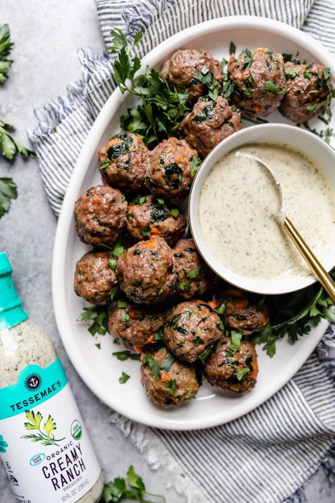 Overhead view of a white platter of the ultimate meal prep meatballs served with a small bowl of ranch dressing on the platter and a bottle of Tessemae's Organic Creamy Ranch Dressing to the side of the platter.