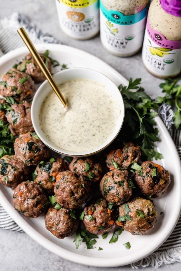 A white platter of the ultimate meal prep meatballs served with a small bowl of ranch dressing with a trio of Tessemae's dressings next to the platter.