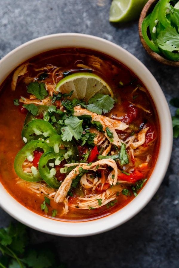 A white bowl on a grey surface filled with Instant Pot Chicken Tortilla-Less Soup that's topped with slices of fresh jalapeños and a lime wedge. 
