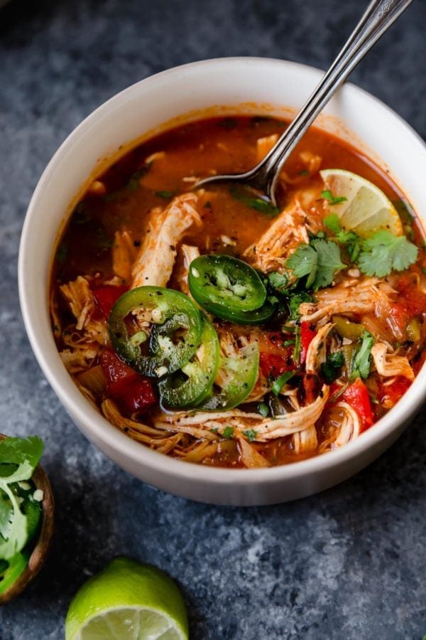 A white bowl on a grey surface filled with Instant Pot Chicken Tortilla-Less Soup. There's a spoon the bowl and the soup is topped with slices of fresh jalapeños and a lime wedge. 